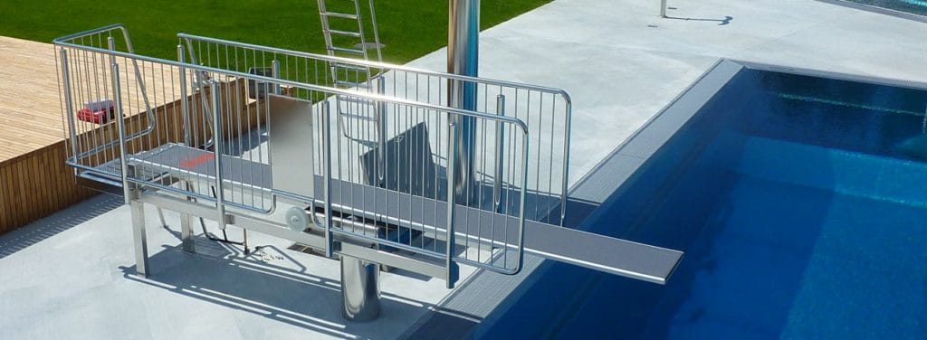 Tremplins : sélection et normes DIN EN 13451-10 ROIGK Ein metallener Pool-Lift mit Geländer befindet sich am Rand eines Schwimmbeckens und ermöglicht einen barrierefreien Zugang. Das Pooldeck ist sauber und geräumig, mit einer Rasenfläche und einem Holzdeck im Hintergrund.