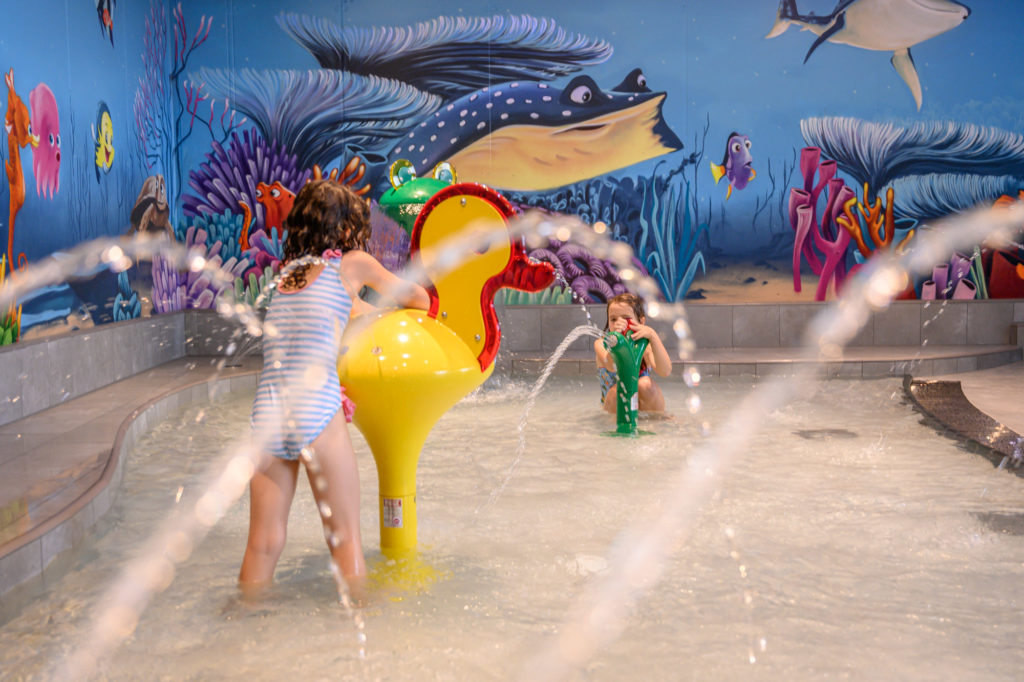 Les thermes du lac de Constance à Überlingen : La nouvelle zone interactive pour les tout-petits ROIGK Zwei Kinder spielen in einem flachen, überdachten Planschbecken mit bunten Wandmalereien zum Thema Meer und spritzen Wasser aus interaktiven Strukturen in Form von Tieren.