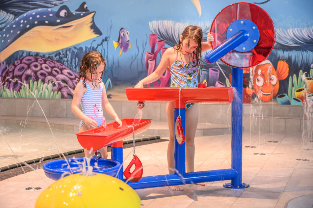 Les thermes du lac de Constance à Überlingen : La nouvelle zone interactive pour les tout-petits ROIGK Zwei junge Mädchen spielen mit interaktiven Wasserspielen in einem Indoor-Wasserzentrum, umgeben von farbenfroher Wandkunst mit Fischen, Korallen und einem Stachelrochen. Um sie herum spritzt Wasser aus verschiedenen Armaturen.