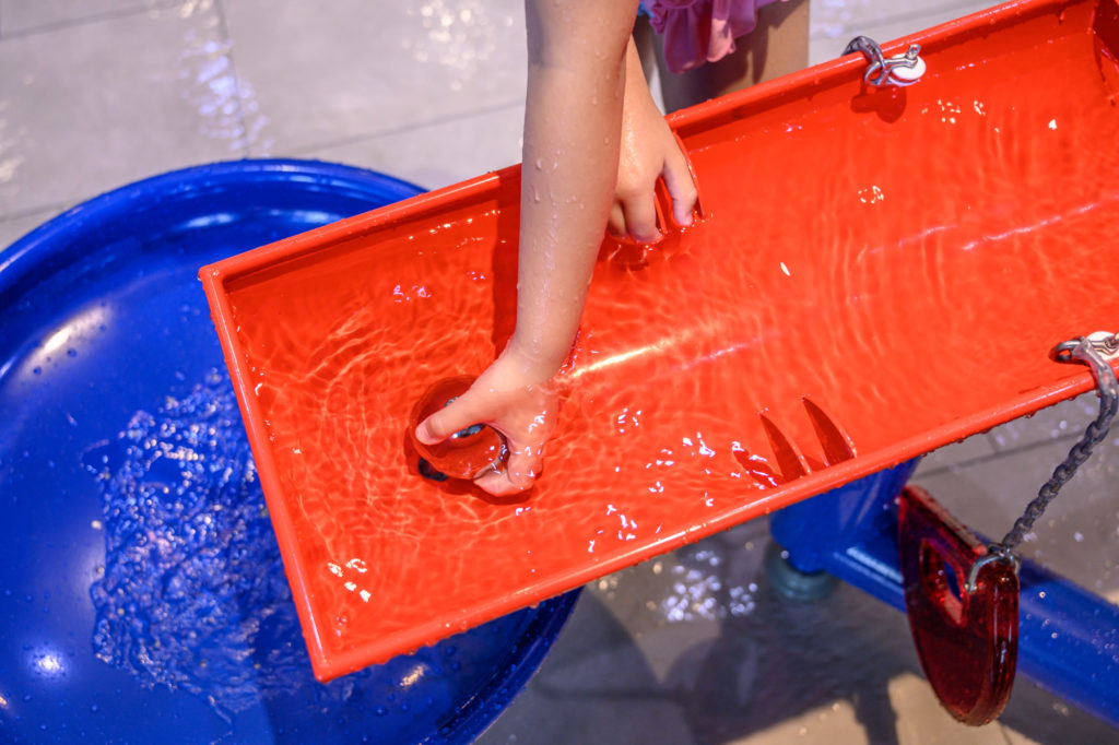 Les thermes du lac de Constance à Überlingen : La nouvelle zone interactive pour les tout-petits ROIGK Die Hand eines Kindes bewegt das Wasser entlang einer roten Kunststoffrinne und lenkt es in ein großes blaues Becken in einem Wasserspielbereich. Die Oberfläche ist nass und die Szene suggeriert interaktives Lernen oder Spielen.