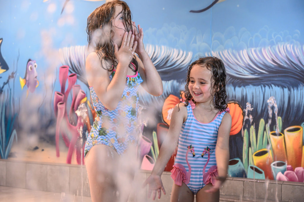 Les thermes du lac de Constance à Überlingen : La nouvelle zone interactive pour les tout-petits ROIGK Zwei junge Mädchen in Badeanzügen spielen lächelnd und lachend an einem Springbrunnen im Haus. Ein farbenfrohes Unterwasser-Motiv mit Fischen, Korallen und Meerespflanzen schmückt die Wand hinter ihnen.