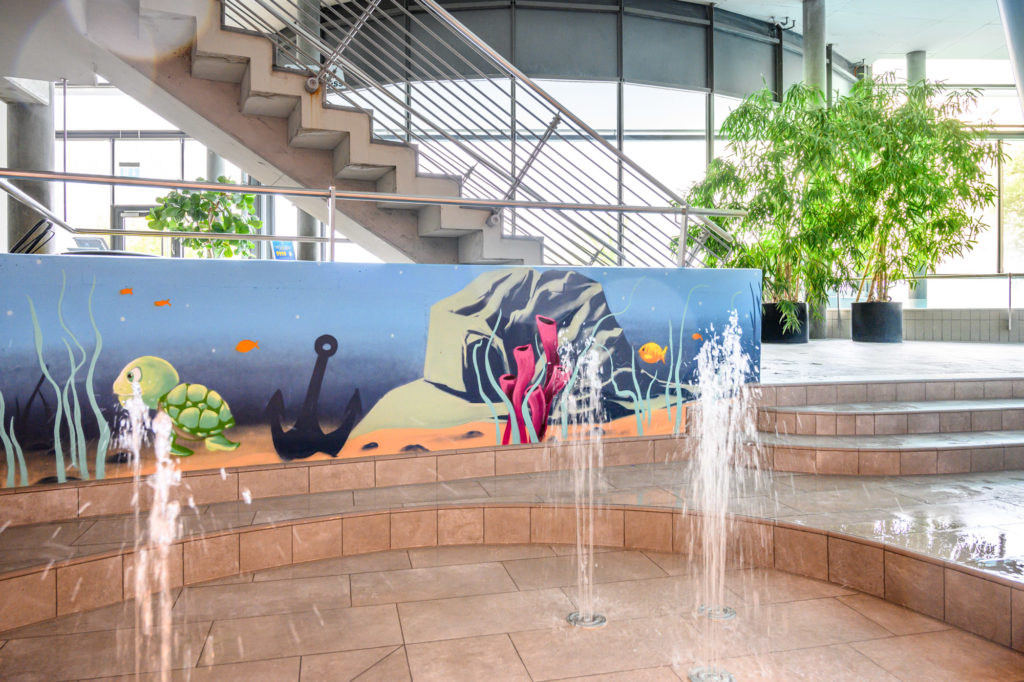 Les thermes du lac de Constance à Überlingen : La nouvelle zone interactive pour les tout-petits ROIGK Ein kleiner Zimmerbrunnen mit Wasserdüsen spritzt aus gefliesten Stufen neben einer Wand mit einer Unterwassermalerei mit einer Schildkröte, Fischen, einem Anker und Felsen; im Hintergrund befinden sich große Grünpflanzen und Treppen.