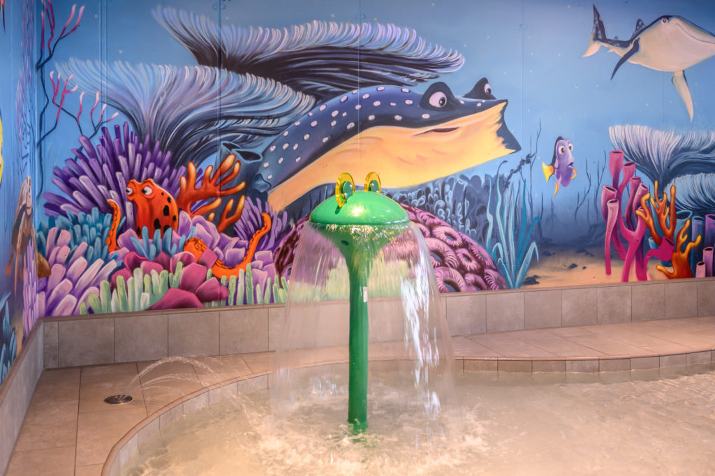 Les thermes du lac de Constance à Überlingen : La nouvelle zone interactive pour les tout-petits ROIGK Ein farbenfrohes, überdachtes Planschbecken mit einer grünen Wasserfontäne und einem Wandgemälde zum Thema Ozean mit Cartoon-Meerestieren, Korallen und Fischen an der Wand dahinter.