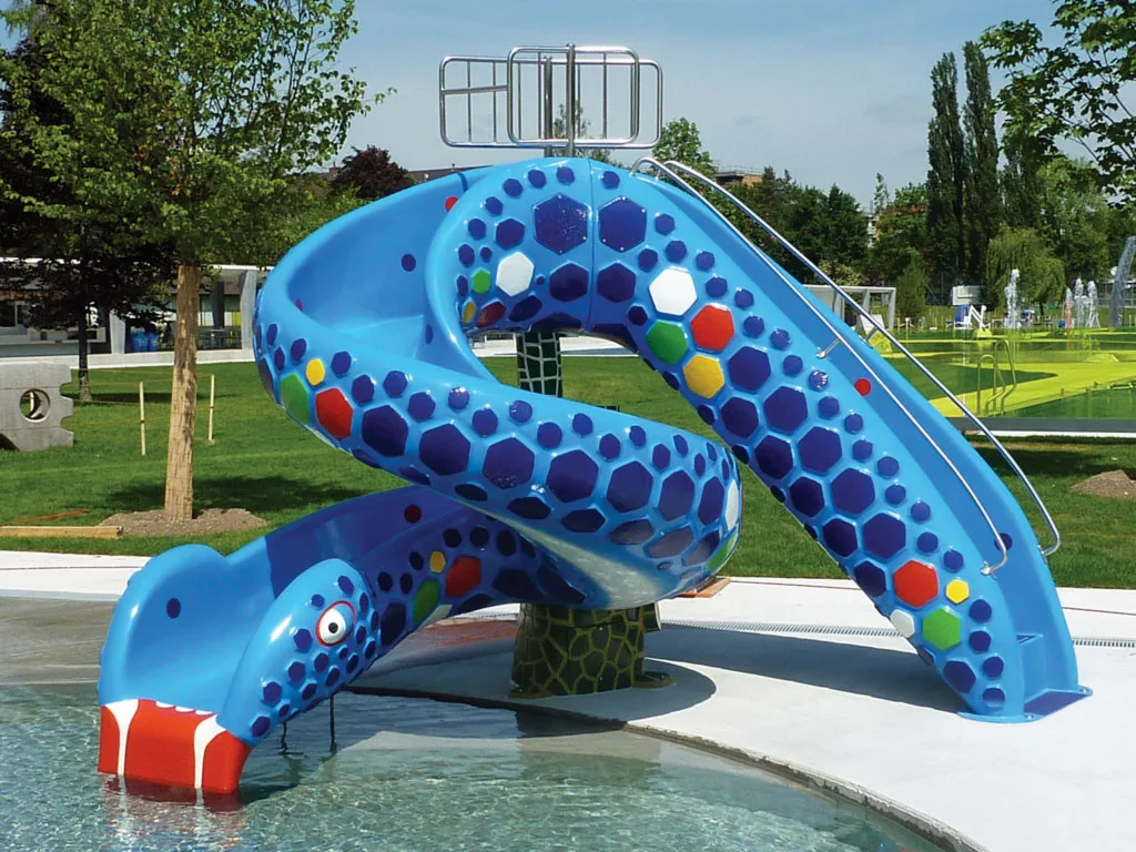 Die Schlangenrutschbahn Kobri R202005-10 ist eine blaue, spiralförmige Wasserrutsche mit sechseckigen Mustern, roten Reißzähnen und großen Augen, die neben einem flachen Außenbecken in einer Parkanlage installiert ist.