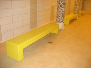 Un espace intérieur carrelé présentant le Banc chaud R200601-3000 sous forme de longs bancs rectangulaires jaunes, avec des colonnes recouvertes de mosaïques, des carreaux de sol beiges et des carreaux muraux crème et beige.