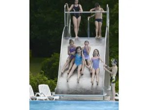 Sechs Kinder in Badeanzügen rutschen die Breite Wasserrutschbahn R150-1BREIT in einem Schwimmbad hinunter, während drei oben warten und einer mit Schwimmbrille unten steht. Weiße Beckenstühle und Grünpflanzen vervollständigen die Hintergrundszene.